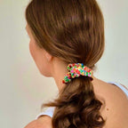 A woman shown from the back wearing a colorful floral fabric scrunchie to hold up her brown hair, set against a plain light beige background.