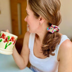 A woman in a white tank top wearing a colorful floral print fabric scrunchie, holding a ceramic mug with a red and green floral pattern.