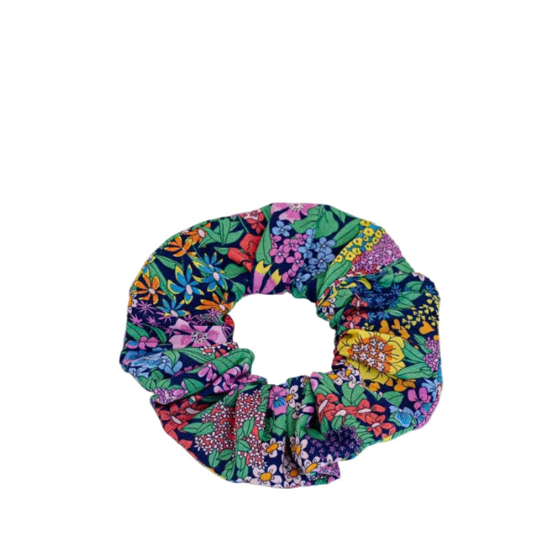 A fabric scrunchie with a dense, colorful floral and botanical print featuring green, pink, purple, blue, and yellow, photographed on a clean white background.